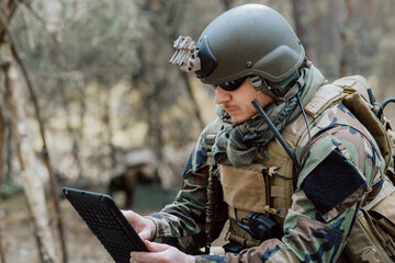 Bearded soldier in uniform sit on military transport crates, analyze data on a tablet and work out...