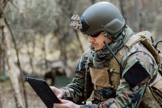 Bearded soldier in uniform sit on military transport crates, analyze data on a tablet and work out tactics at a temporary forest base. In the background, you can see a soldier protecting the base.