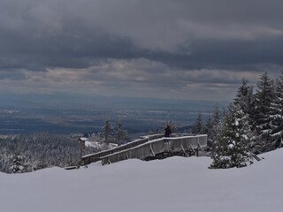 Hugging couple enjoys the stunning panoramic view from a wooden observation deck near Schliffkopf, Germany in Black Forest with Rhine valley, Strasbourg and Vosges on horizon in winter season.
