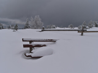 Wooden rest bench covered by deep snow in front of fence and frozen trees on hiking tour near Schliffkopf hill, Germany in Black Forest mountain range on cloudy winter day.