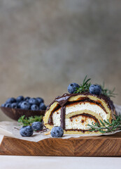 Sponge cake roll with chocolate and cream cheese decorated with chocolate glaze, blueberry and rosemary on parchment, light background. Biscuit swiss roll.