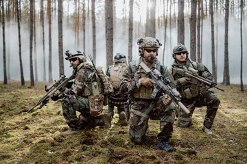 Poster Four fully equipped, middle-aged soldiers in camouflage uniforms form a line, ready to fire, aiming with their rifles. A military operation in action, a unit standing in a dense forest. © ABCreative