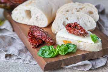 Sliced ciabatta bread with sun-dried tomatoes, cheese, basil and olive oil on wooden board, stone background.