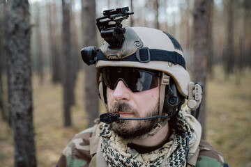 Portrait of a middle-aged bearded soldier in a Woodland military uniform and helmet, with headphones on his head.