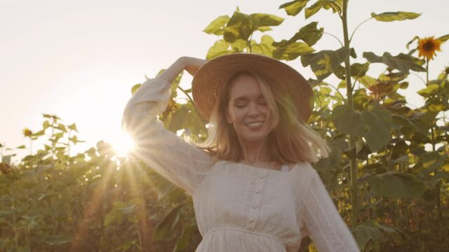 Medium slowmo close up of romantic young woman in summer dress and straw hat twirling while walking along rural sunflower field at sunset