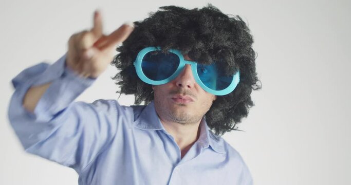 Man with curly hairy wearing funny and big sunglasses dancing showing victory sign isolated on gray background. Party, music, lifestyle, radio and disco concept. 