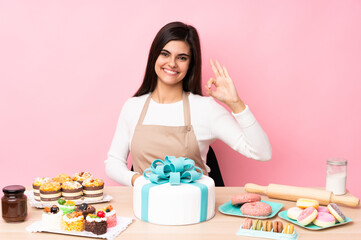 Obraz na płótnie Canvas Pastry chef with a big cake in a table over isolated pink background showing ok sign with fingers