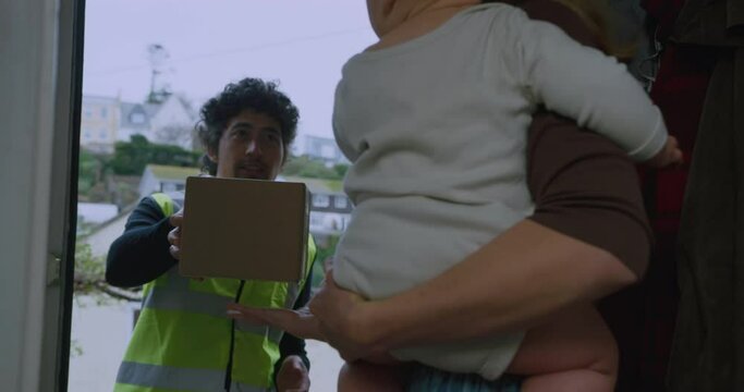 A young mother is taking a delivery at her front door while holding her baby