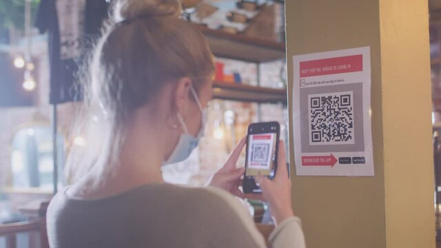 Woman wearing mask with mobile phone checking into venue scanning QR code during health pandemic - shot in slow motion