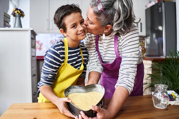 Caucasian mother with gray hair gives a kiss to her son showing the heart cake they have prepared...