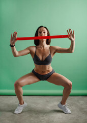 A woman in sportswear is engaged in fitness. She lifts swings her bicep with a red elastic band on a green background