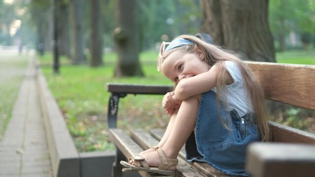 Small happy child girl sitting on a bench resting in summer park.