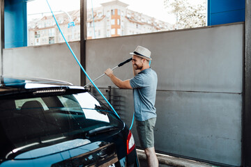 Young man with hat washing his car during daylight at car wash station using high pressure water.