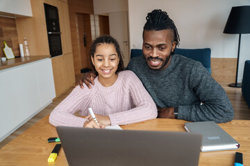Happy smiling African American father helps to study online to his teen age nice daughter. E...