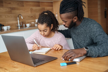 Focused African American father helps to study online his teen age nice daughter. E-learning distant studying online at home. Family doing homework, watching online course, webinar with teacher in app