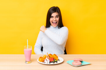 Young woman eating waffles and milkshake in a table over isolated yellow background celebrating a victory