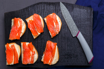 Bruschettes with butter and salmon on a black wooden board on a gray background near a knife.