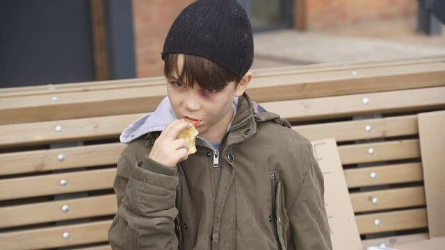 Sad street boy is eating food on street, want to be loved and have shelter, portrait of unhappy child in dirty clothes.