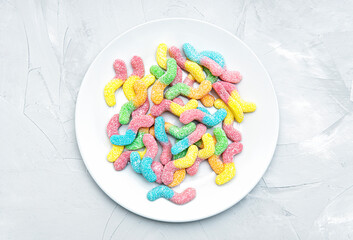 Top view of a white plate full of colored sugar coated gummy worms on a concrete table