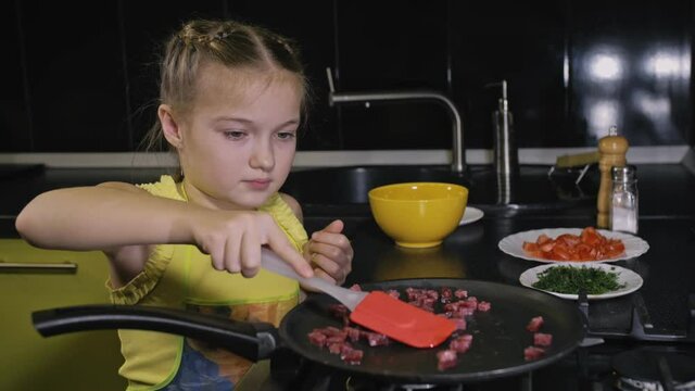 Smart girl learning to cook. Happy and serious young mistress children to cook a Neapolitan egg fried omelette from sausage. Modern Built In Kitchen Appliances in green black color.