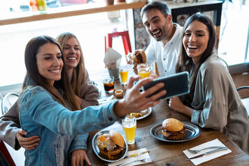 Happy group of friends taking selfie with smart phone while eating burgers in a restaurant.