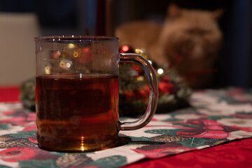 View of a cup of tea on a christmas background with an orange cat. Transparent glass with tea on a red table with ornaments.