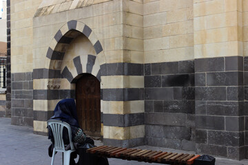 Old woman with local clothes sitting on a chair in the street in front of a historic building in Gaziantep, Turkey.
