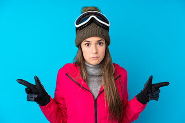 Teenager skier girl with snowboarding glasses over isolated blue background pointing to the laterals having doubts