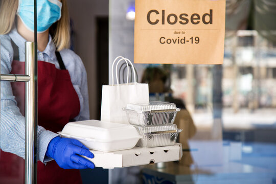 Woman Waiter In Protective Medical Mask And Gloves Work With Takeaway Orders. Waiter Giving Takeout Meal While Lockdown, Coronavirus Shutdown. Food For Delivery Against Sign Closed Due To Covid 19