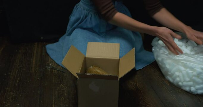 A young woman is wrapping up a vase in a box with styrofoam balls to protect it