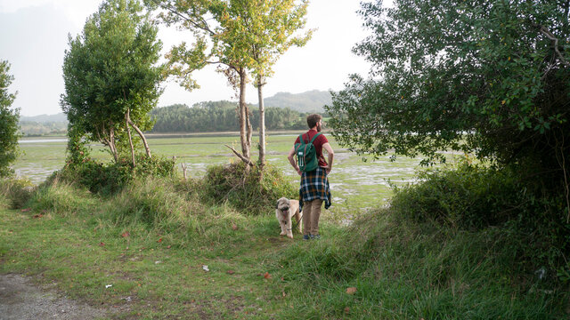 Hombre joven barbudo con mochila y su perro contemplando paisaje de humedal
