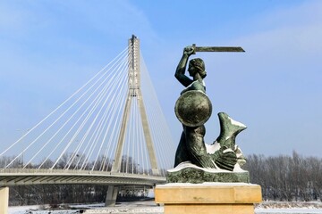 Fototapeta na wymiar Statue of Mermaid (Polish: Warszawska Syrenka) symbol of Warsaw by the Vistula river on the background of Swietokrzyski Bridge. Winter time in Warsaw, Poland. 