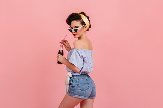 Slim pinup girl in denim shorts looking over shoulder. Studio shot of beautiful young woman with bottle of soda standing on pink background.