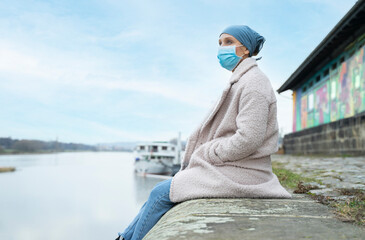 Young woman with cancer, wearing a blue bandana, sitting on a small wall above the river, with a thoughtful expression.