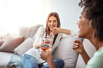 Happy young female friends with wine on sofa at home. Happy smiling young women friends opening boottle of wine at home. Girls havin fun. Two Female Friends Relaxing With Glass Of Wine 