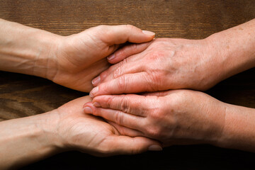 Old mother hands in daughter palms on dark wooden table background. Lovely, emotional, sentimental moment. Closeup. Concept of care about parent.