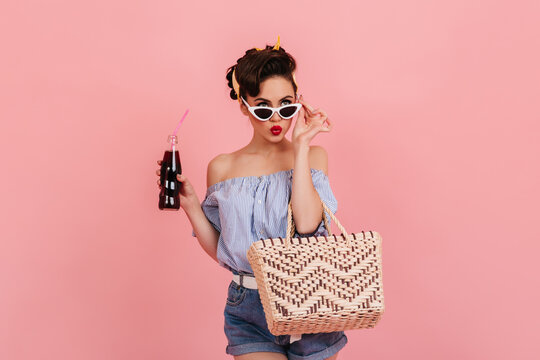 Pensive pinup girl with bottle of soda touching sunglasses. Studio shot of refined elegant woman isolated on pink background.