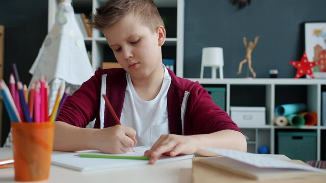 Serious child is drawing with colorful pencils sitting at table in apartment enjoying creative activity concentrated on art. Childhood and leisure time concept.