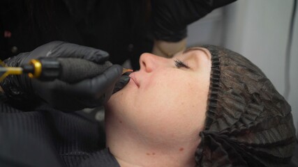 The procedure of permanent lip makeup in the beauty salon. Cosmetologist pricks paint on the lips of a young girl with a microneedle. The process of applying permanent makeup to the lips.