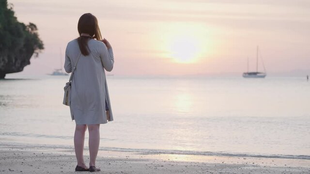 Young asian woman enjoying moment during sunset at the island beach, using smartphone taking pictures of beautiful ocean scenic, tropical island travel destination, single woman lone journey