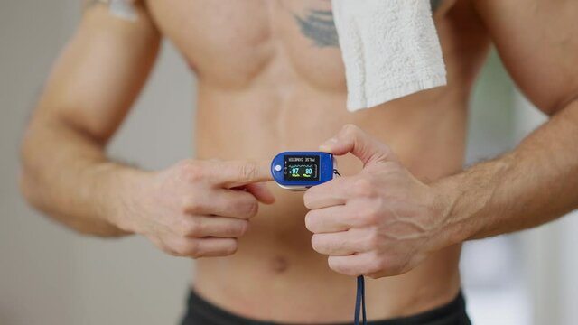 Unrecognizable young sportive man measuring oxygen saturation with pulse oximeter. Athletic muscular sportsman controls pulse rate and perfusion index training indoors. Healthy lifestyle concept.