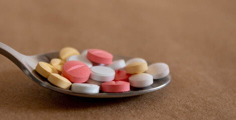 Close up of a spoon full of pink, white and yellow tablets on light brown background
