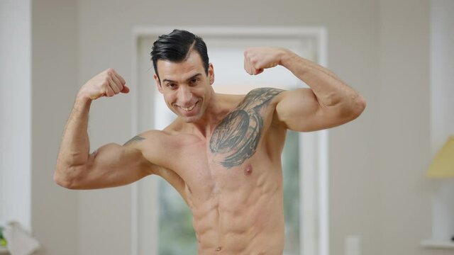 Tattooed confident sportsman pointing at camera smiling and raising hands showing muscles. Portrait of young handsome Middle Eastern man bragging biceps indoors posing at home.