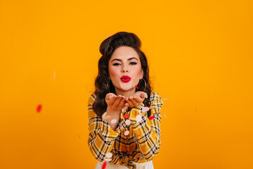 Brunette pinup girl posing with kissing face expression. Amazing young lady in checkered shirt standing on yellow background.
