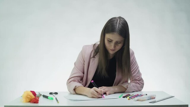 Attractive young woman uses a pink marker to draw something on on a white paper at the table in a white studio