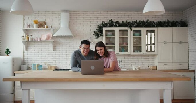 Cheerful young tanned man and woman surf internet using contemporary laptop at wooden table in light kitchen at home slow motion