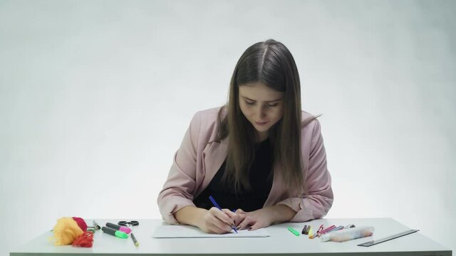 Attractive young woman uses markers to draw picture on on a white paper at the table in a white studio