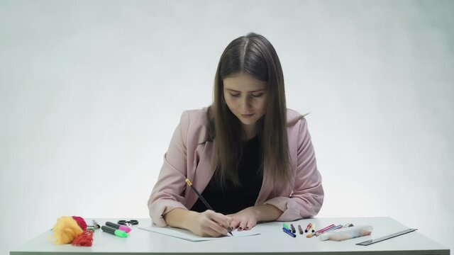 Attractive young woman uses pencil to write down something on on a white paper at the table in a white studio
