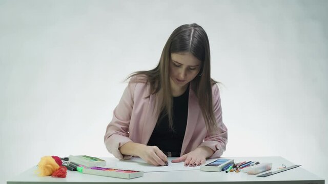 Attractive young woman uses pastel to draw something on on a white paper at the table in a white studio