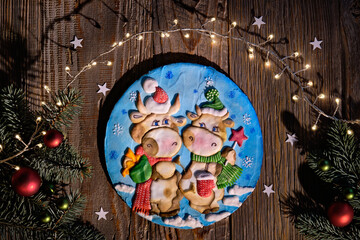 Decorated gingerbread with two cartoon cow characters. Christmas flat lay with fir twigs, red baubles and stars confetti. Dark wooden planks, top view of rustic wooden table.